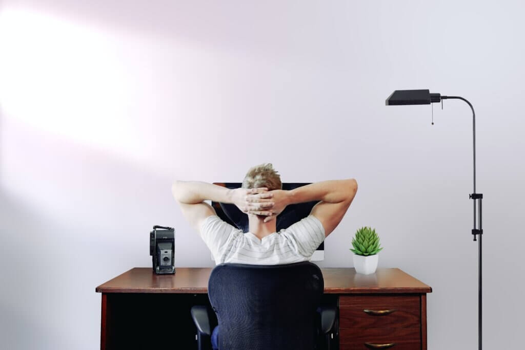 Man sitting at computer while working from home, a desk plant is on his right and a old-fashioned camera is on his left. His hand are crossed over the back of his head.