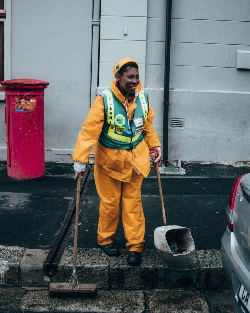 Cleaner in Orange Overalls holding dustban and broom as he cleans.